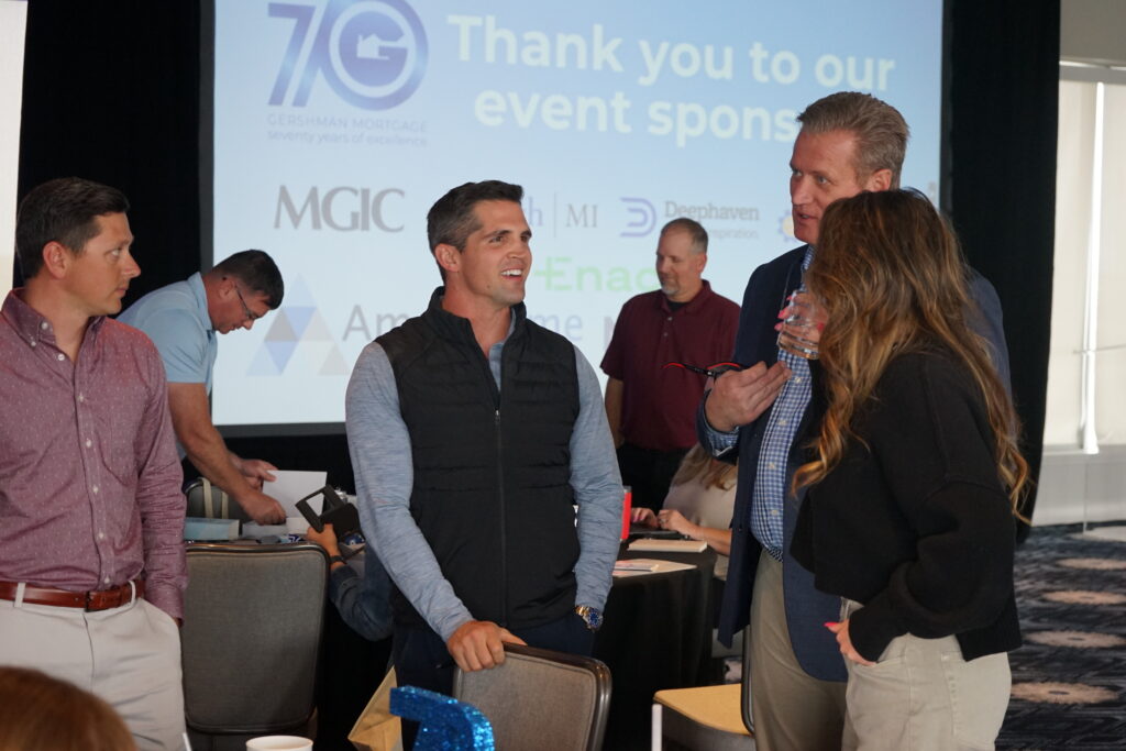 A group of people standing together talking at a corporate event, with branded signage visible.
