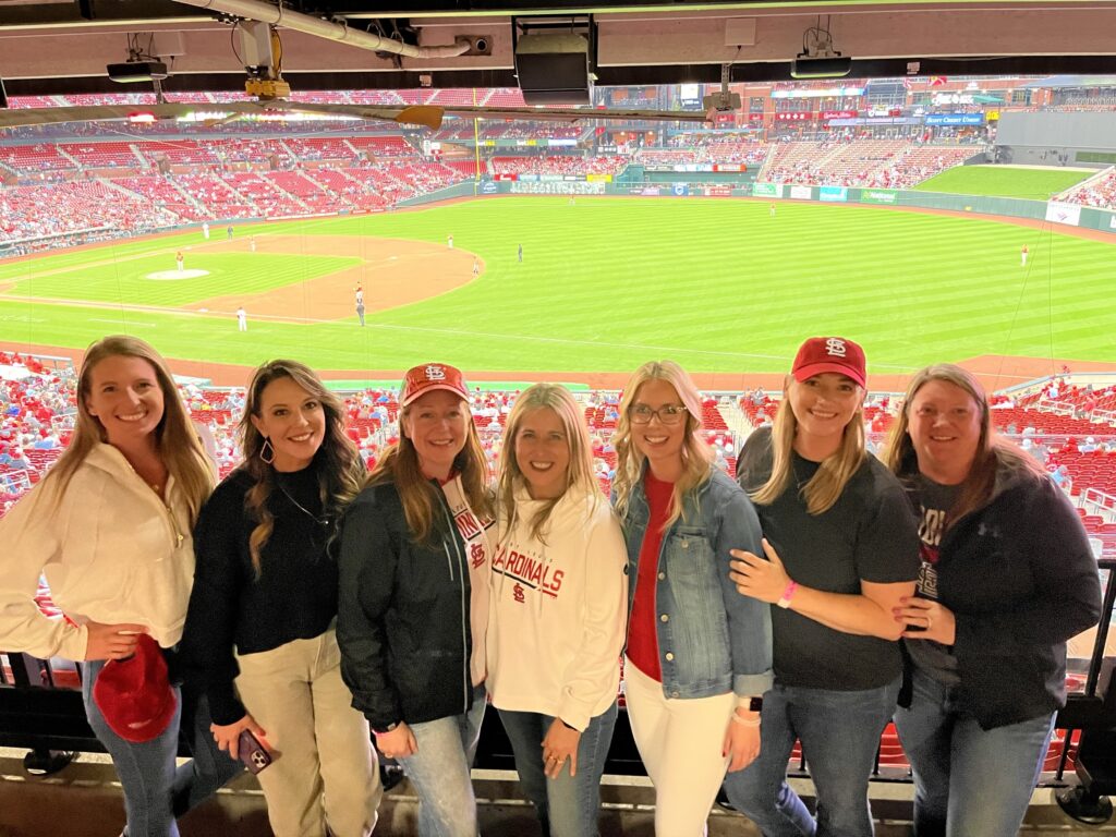 A group of seven women posed in front of the field at Busch Stadium during a St. Louis Cardinals game.